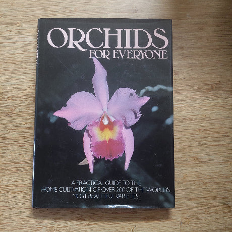 Book titled 'Orchids for Everyone' with a pink orchid on the cover, placed on a wooden surface.